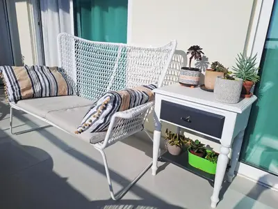 a white wicker chair and a nightstand with plants on a balcony at Sea View Floral Suite - 2 bedroom in Larnaka