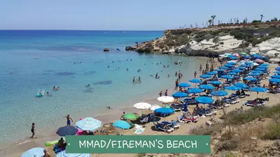 a beach with blue and white umbrellas and people in the water at CHRIS'S KYKLADES HOLIDAY APARTMENT in Paralimni