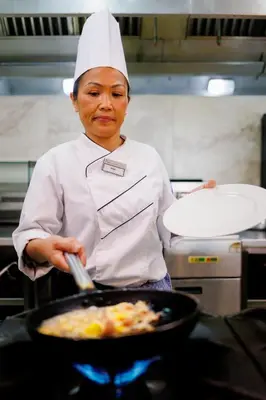 a woman in a kitchen preparing food in a pan at Tasia Maris Beach Hotel - Adults Only in Ayia Napa